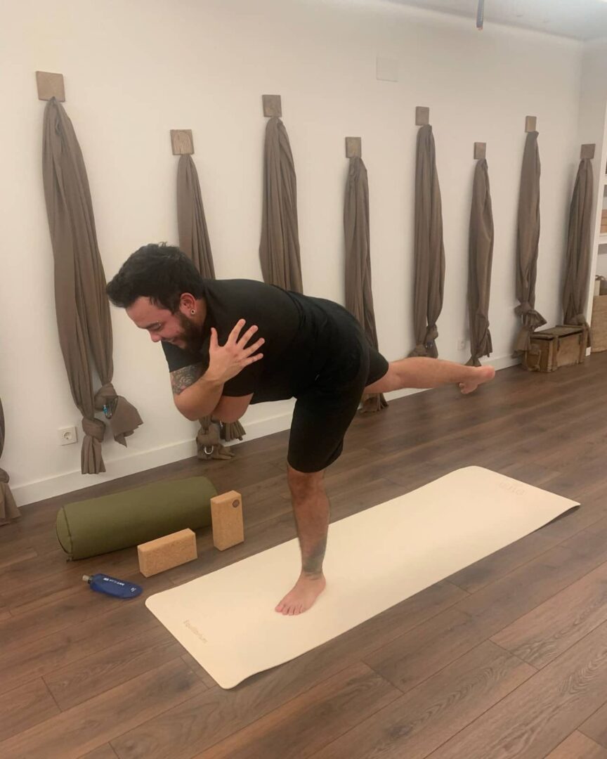 A man in a black outfit balances in Warrior III pose on a beige yoga mat, arms crossed over his chest, inside a yoga studio with aerial silks hanging on the wall. Yoga props including a bolster, two cork blocks, and a water bottle are neatly placed beside the mat on a wooden floor.