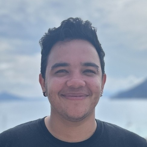 Smiling man with short, dark curly hair and facial piercings, wearing a black shirt, standing in front of a soft-focus coastal background with mountains and a cloudy sky.