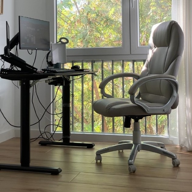 Bright home office with a beige office chair beside a black standing desk, dual monitors, and a tangle of cords underneath. Sunlight filters through large windows.